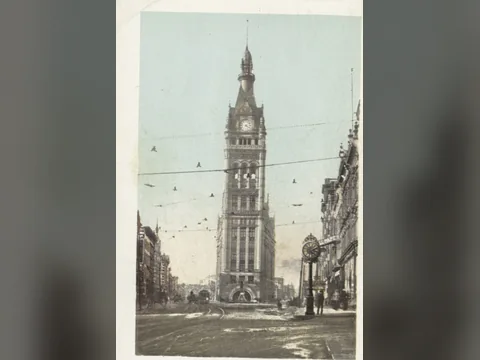 Postcard looking straight down a street at the narrow end of City Hall: a tall stone clock tower with a pointed top, overhead wires and a street clock.