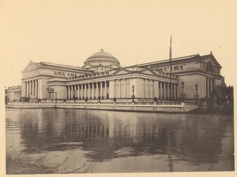 Black and white 1893 photo of the Art Palace across water: a low dome over long colonnades and pedimented pavilions, reflected in the lagoon.