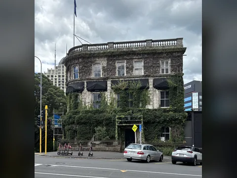 Colour photograph of a three-storey plastered building almost covered in creeper, with a balustrade along the roof and parked cars and hire scooters in front.