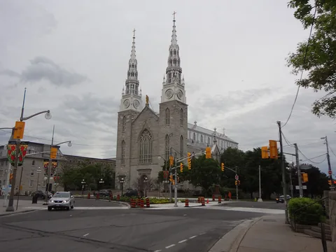 A grey stone Gothic church front with twin silver spires, a long nave running back to the right, a street junction with traffic lights in the foreground.