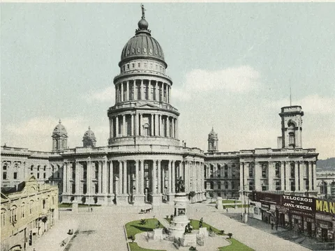 Postcard of a pale domed hall facing a plaza: a tall colonnaded drum and dome, long classical wings with corner towers, and a monument in a small garden.