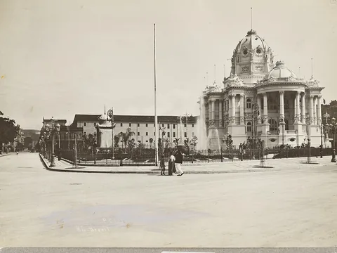 A domed white palace with columned porticos and fountains beside a wide empty avenue; a couple stand by a railing, and the print is mounted on grey card.