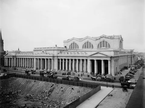 Black and white photo of the near-finished station from above a street: a long Doric colonnade, a pedimented entrance, great arched windows, horse carts below.