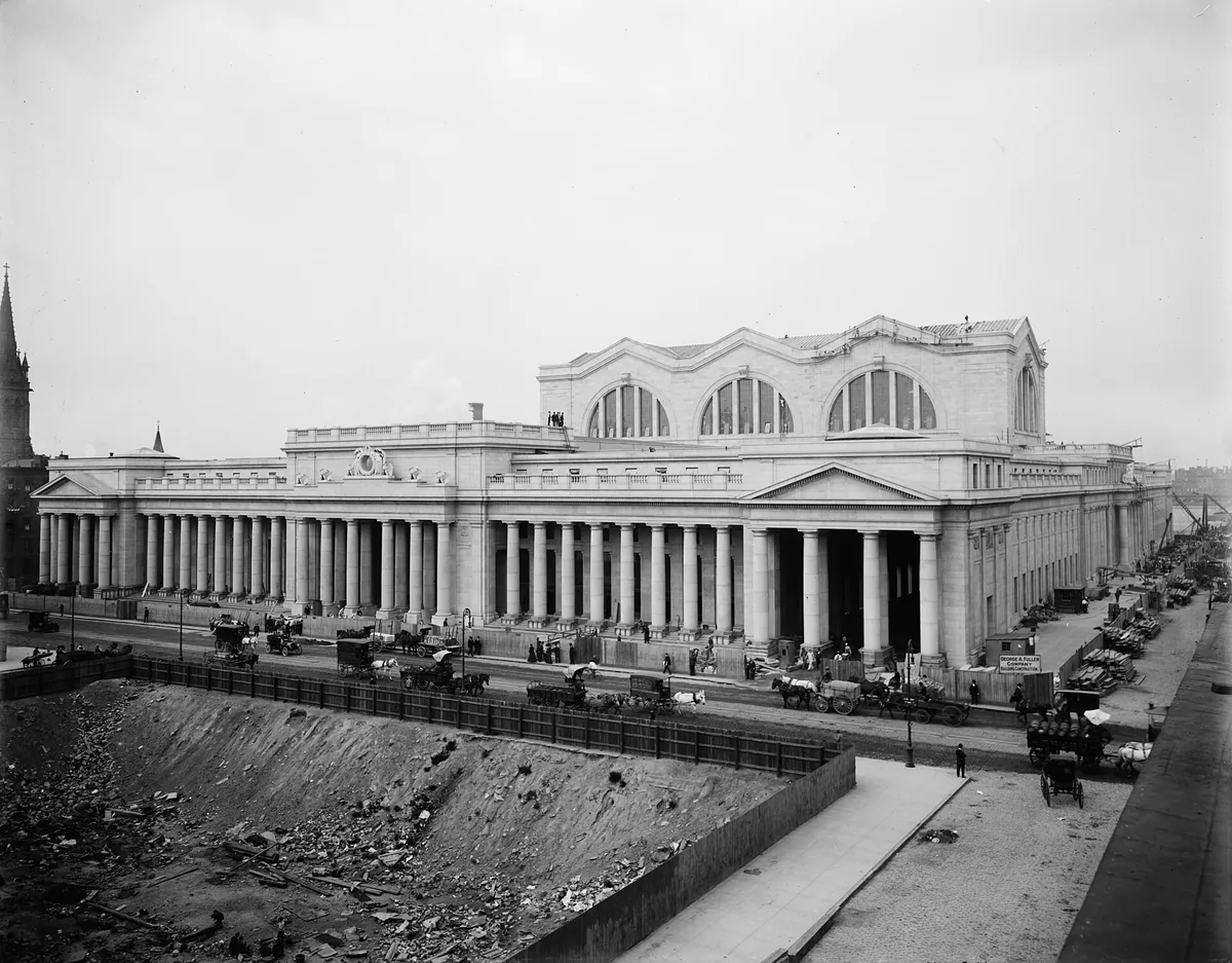 A long classical stone station with a row of columns, seen from above a wide street with a cleared site in front.