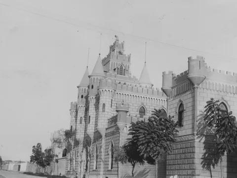 A crenellated castle with pointed turrets, seen at an angle along a dusty street, with young trees along its wall and overhead wires.