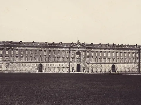Sepia photograph of an immensely long, flat-fronted palace with rows of identical windows and three arched gateways, seen across an empty dark field.
