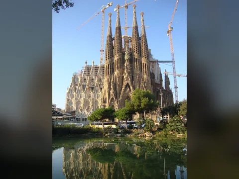 Colour photograph of a huge unfinished church with four tapering stone towers and construction cranes, reflected in a pond among trees.