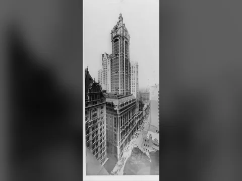 Black and white photo looking down on the tower from high up: a narrow shaft with a domed lantern top rising over a mansard-roofed block and a crowded street.
