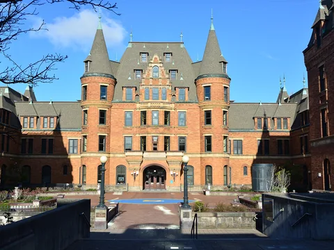 A wide brick chateau-style school with two conical towers and a steep slate roof, seen straight on across a paved forecourt with lamp posts.
