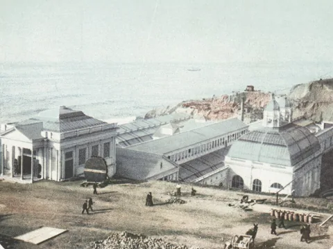 Postcard of the seaside complex from the hill: a Greek-fronted entrance, long glass roofs, a domed glass hall, figures on bare ground and the Pacific beyond.