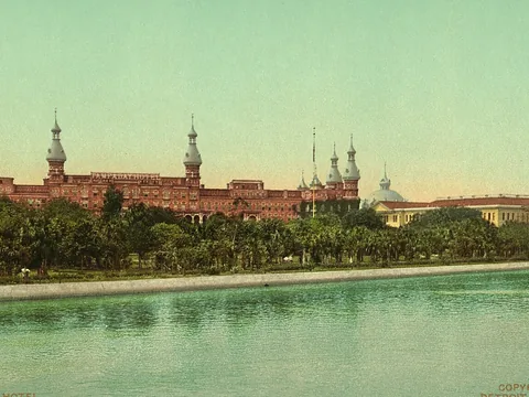 Colour print of the long red brick hotel across the river: onion-domed minarets rising above a line of palms, with green water in front and a tall chimney at right.