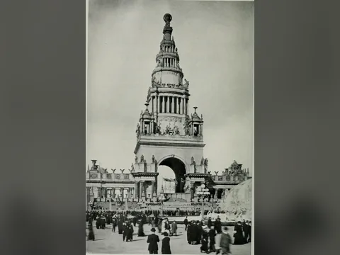 Black and white photo of the tiered tower over a great arch, statues on its ledges, a globe on top, and a crowd in dark coats in the court in front.