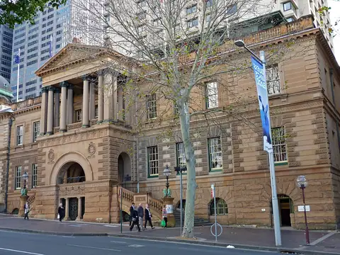 Colour photograph of a sandstone building with a columned portico above a rusticated arched entrance, a bare tree in front and tower blocks behind.