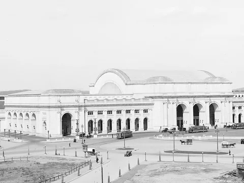 Black and white photo of the white marble station from high up: three great entrance arches, a vaulted hall behind, trolleys on the plaza and bare ground in front.