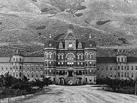 A long symmetrical brick asylum with a domed central block, clock tower and end pavilions, at the foot of bare hills, with a dirt drive and young trees in front.