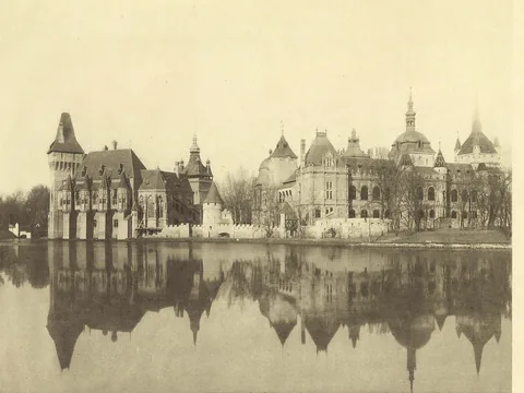 Sepia photograph of a long group of castle towers, spires and domes along a still lake, the whole group mirrored in the water.
