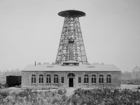A long low brick laboratory with arched windows, and behind it a tall timber lattice tower topped by a wide disc-shaped frame, on cleared scrub land.