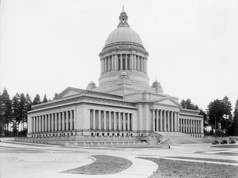 Black and white photo of the new domed Legislative Building at an angle: tall ribbed dome on a colonnaded drum, columned wings, young trees and a few parked cars.