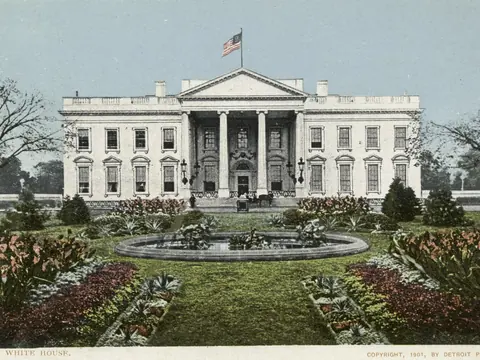Postcard of the north front: a white house with a columned portico and a flag on the roof, behind a round fountain, a lawn and beds of flowers.
