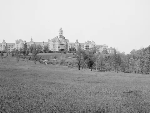 A long hospital complex with a central clock tower and stepped wings on a rise, seen far off across a sloping meadow with trees.